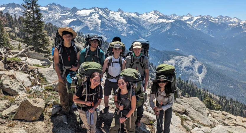 A group of people wearing backpacks stand on rocky ground, high above a snow covered mountainous landscape. 